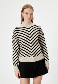 Sweter w paski w jasnym beżu i czerni, z wysokim kołnierzem, luźnymi rękawami i wzorem w zygzak. Nosi się go z czarną plisowaną spódnicą.