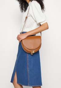 Bolso de cuero marrón tipo bandolera con forma curvada de media luna y herrajes dorados. Presenta un charm decorativo y una larga correa ajustable.