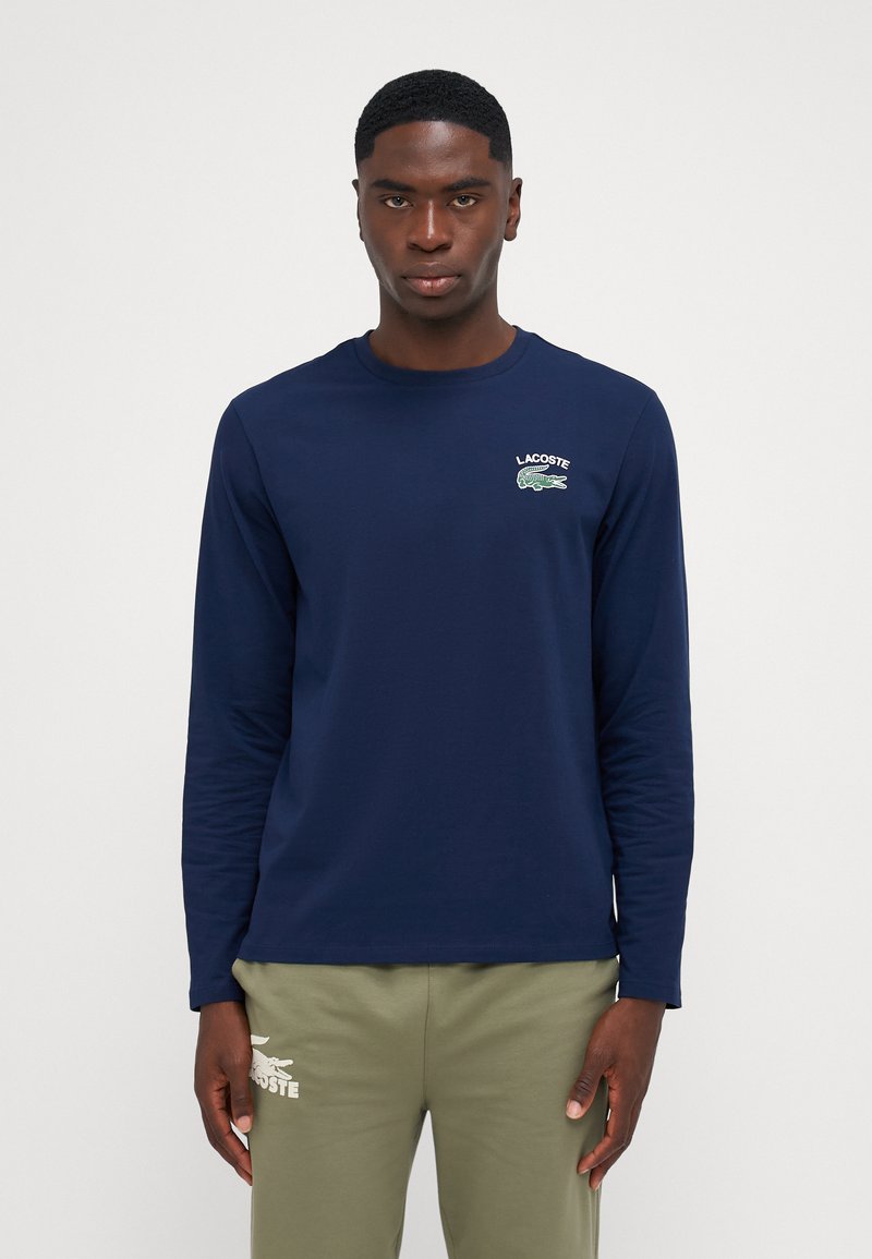 Chemise à manches longues marine avec encolure ronde, arborant un logo de crocodile vert et le texte "LACOSTE" en blanc. Tissu lisse et coupe ajustée.