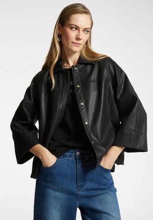 Giacca di pelle nera con colletto alto e bottoni a scatto. Presenta maniche ampie e una superficie liscia, abbinata a jeans in denim blu.