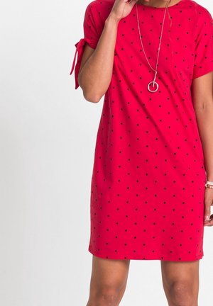 Femme portant une robe rouge mi-longue à pois noirs, manches courtes nouées en nœuds, et un long collier avec un pendentif circulaire.