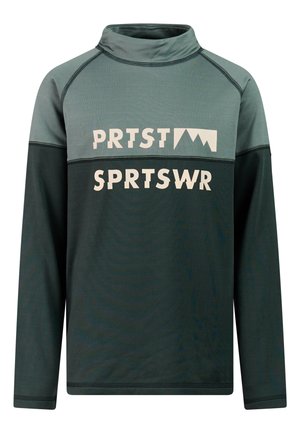Jongen staat met gevouwen handen, draagt een groen en blauwgroen lang mouw shirt met tekst en losse groene broek tegen een effen achtergrond.