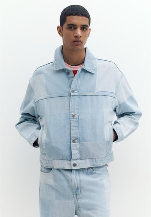 Tânăr purtând o jachetă din denim patchwork albastru deschis și blugi asortați, stând cu mâinile în buzunare, pe un fundal alb.