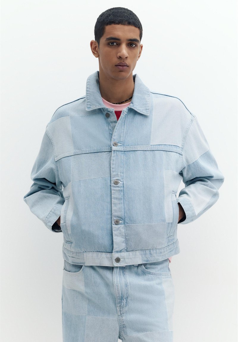 Jeune homme portant une veste en jean patchwork bleu clair et un jean assorti, debout, mains dans les poches, sur fond blanc.