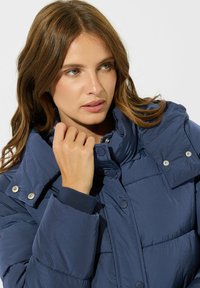 Chaqueta acolchada azul marino con cuello alto, botones a presión y puños elásticos. Textura suave con detalles de costura en toda la prenda.