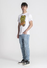 Camiseta blanca de algodón con un gráfico de oso colorido goteando. Combinada con jeans azul claro y zapatillas negras con detalles blancos.