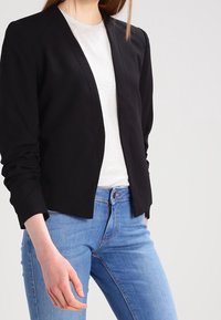 Černé sako s výrazným střihem, dlouhými rukávy a měkkým materiálem, nošené přes světle šedé tričko a kombinované s modrými džínami.