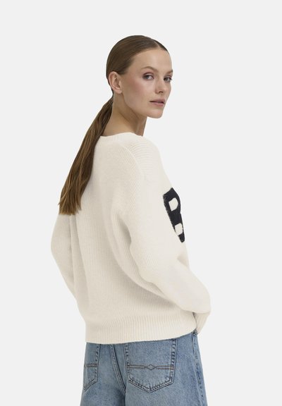 Pull en maille blanche avec un col rond et des coutures d'épaule abaissées. Présente une grande lettre noire "B" sur le devant. Associé à un jean en denim bleu.
