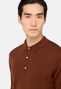 Rudas mezgimo polo marškinėliai su mygtuku ant apykaklės, kuriuose yra trys šviesios spalvos mygtukai ir tekstūruota medžiaga.