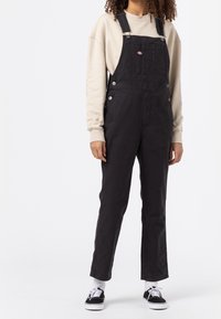 Zwarte denim salopettes met verstelbare banden, een voorzak en zilverkleurige hardware, gedragen over een lichtbeige sweatshirt en zwarte sneakers.
