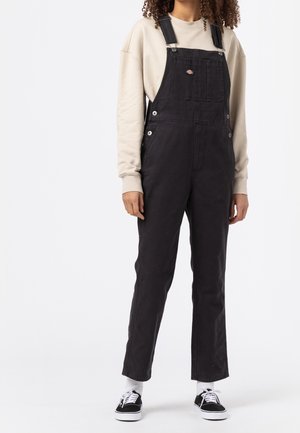 Salopette in denim nero con spalline regolabili, tasca frontale e dettagli in metallo argentato, indossata su una felpa beige chiaro e sneakers nere.