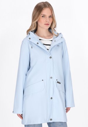 Kobieta ubrana w jasnoniebieską pelerynę przeciwdeszczową z kapturem i zatrzaskami, na czarno-białej koszulce w paski i niebieskich dżinsach.