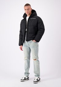 Zwarte pufferjack met een capuchon, voorzien van een rits aan de voorkant en zijzakken. Draagt over een wit shirt, gecombineerd met versleten lichtblauwe spijkerbroeken.