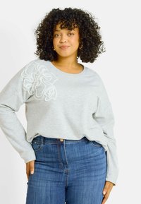 Sudadera gris claro de manga larga, con bordado floral blanco texturizado en el hombro izquierdo, combinada con vaqueros de cintura alta azules.