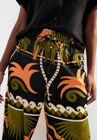 Pantalones holgados de tela negra con un estampado tropical en tonos verdes, naranjas y crema; cinturilla elástica con cordón ajustable.
