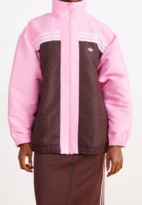 Jacka med dragkedja med en rosa övre del och en brun mönstrad nedre del, prydd med vita ränder och en broderad logotyp på bröstet. Elastiska ärmslut.