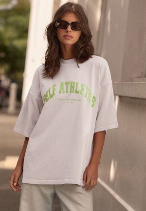T-shirt bianco oversize con la scritta "SELF ATHLETICS" in testo verde brillante. Tessuto morbido, vestibilità rilassata, maniche corte. Abbinato a pantaloni grigi chiari.