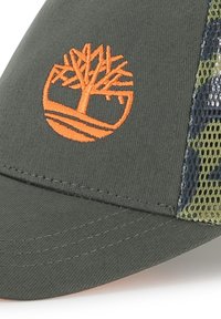 Casquette verte avec un logo d'arbre orange bien en vue. Les panneaux latéraux en mesh ajoutent de la texture et respirabilité. Le design à visière plate renforce sa forme.