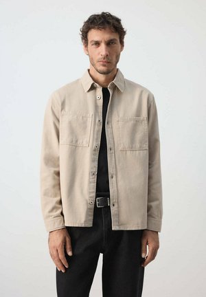 Mann mit lockigem Haar trägt ein beiges Hemd mit Knöpfen über einem schwarzen Top und dunkler Hose vor einem einfarbigen hellen Hintergrund.
