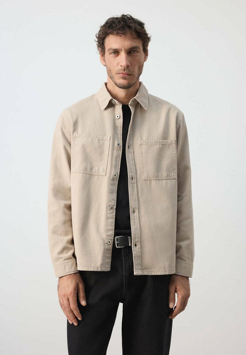 Homme aux cheveux bouclés portant une chemise beige boutonnée sur un top noir et un pantalon foncé sur un fond clair uni.