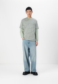 Camiseta a rayas en verde y blanco con mangas cortas superpuesta a una camiseta de manga larga verde, combinada con unos vaqueros ligeros y holgados en azul claro y zapatos negros.