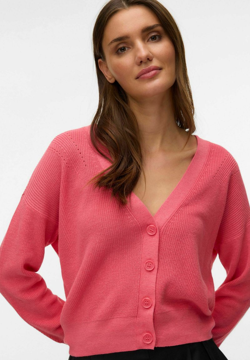 Mujer de cabello castaño que lleva un cárdigan rosa con botones y cuello en V, posando frente a un fondo claro y liso.