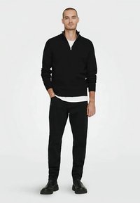 Musta vetoketjullinen neule, resoriset hihansuut, yhdistetty mustiin housuihin. Malli seisoo neutraalissa asennossa, jalassa mustat kengät ja taustana yksivärinen tausta.