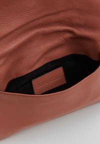 Brązowa skórzana torebka o miękkim, teksturowanym wykończeniu. Wnętrze podszewkowane jest czarnym materiałem, a na nim znajduje się brązowa skórzana metka z logo.