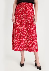 Jupe fleurie rouge avec une taille smockée, de longueur genou, ornée de petites marguerites blanches et d'un tissu léger. Associée à des escarpins noirs.