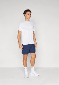 Mees, kes seisab tühja seina vastu, kannab valget sportlikku särk, tumesiniseid lühikesi pükse, valgeid Nike sokke ja valgeid sneakers'e, vaatab otse ette.