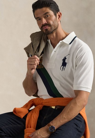 Άνδρας που φοράει λευκό πόλο με μπλε και πράσινη ρίγα και λογότυπο παίκτη πόλο, σκούρα τζιν, πορτοκαλί πουλόβερ δεμένο στη μέση, κρατώντας καφέ μπουφάν στον ώμο.