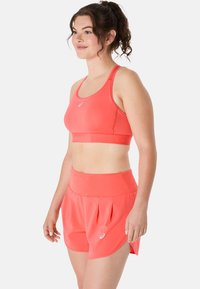 Bright coral sports bra with thin straps and a branded band; matching pleated shorts with a wide waistband and side slits.