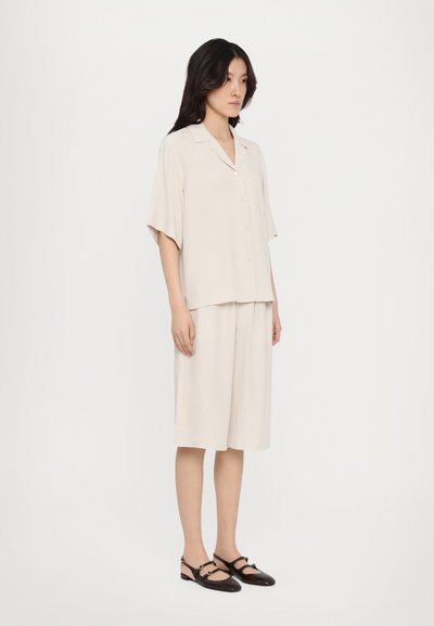 Lys beige kortærmet skjorte med knapper parret med matchende knælang shorts. Glat stof, afslappet pasform og subtil glans. Sorte flade sko.