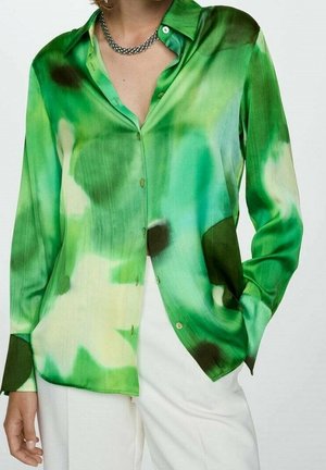 Kobieta ubrana w zielono-białą bluzkę z długim rękawem i guzikami w stylu tie-dye, połączoną z białymi spodniami oraz srebrnym łańcuszkiem na szyję.