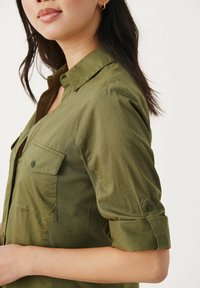 Camicia verde oliva con colletto e polsini abbottonati, caratterizzata da tasche frontali e un pannello laterale testurizzato. Tessuto leggero con una finitura liscia.