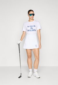 Fehér golf szoknya és környakú póló, tengerészkék szöveggel: "le CLUB du MALBÔN." Kiegészítők között kesztyű és oversized tükröződő napszemüveg található.