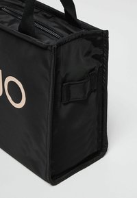 Borsa a tote in nylon nero con forma rettangolare, dotata di zip superiore, manici intrecciati corti e una texture liscia. Dettaglio del logo ben visibile.
