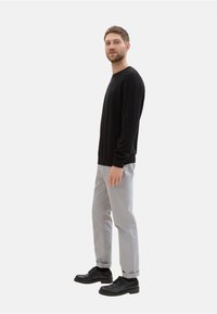 Schwarzer Langarmpullover, hellgraue Hose und schwarze Schuhe. Der Pullover hat einen Rundhalsausschnitt; die Hose ist leicht tailliert mit hochgekrempelten Säumen.