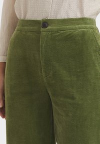 Green corduroy pants with vertical ribbing, featuring a black button at the waist and a smooth texture. Close-up view of the fabric.