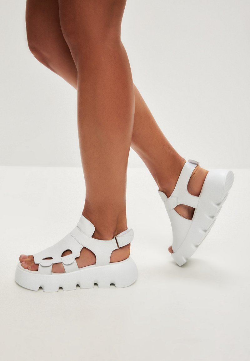 White sandals with a chunky sole and open-toe design. Made of smooth material, featuring cutouts and adjustable ankle strap.