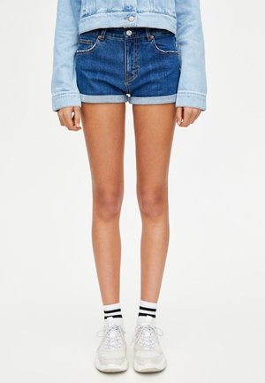 Person iført mørkeblå optrukne denimshorts, lyseblå denimjakke, hvide sneakers og hvide sokker med to sorte striber.