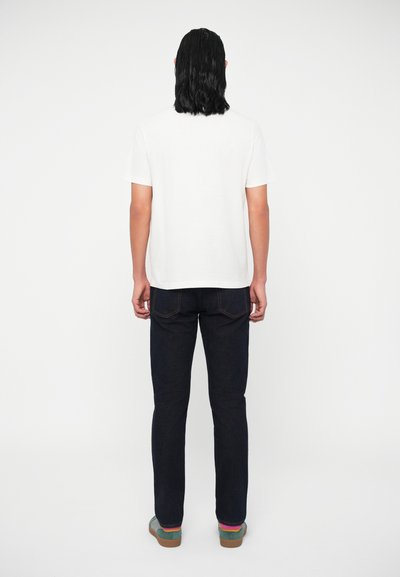 Asmuo su pečių ilgio tamsiais plaukais, vilkintis paprastą baltą marškinėlį, tamsias džinsines kelnes ir mėtinių spalvos sportinius batelius, stovi nugara pasukęs baltame fone.