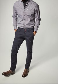 Man som bär en grå knappnupen skjorta, svart polotröja, smala mörkblå byxor och bruna läderstövlar, står med händerna i fickorna.