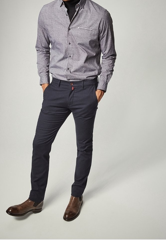 Man som bär en grå knappnupen skjorta, svart polotröja, smala mörkblå byxor och bruna läderstövlar, står med händerna i fickorna.