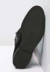 Czarny zamszowy but z widocznym zamkiem błyskawicznym, podeszwa skierowana do przodu, oparty o biały mur na białej powierzchni.