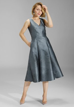 Szara satynowa sukienka w kształcie litery A z dekoltem w kształcie V, bez rękawów, w kwiatowy wzór, z dwiema bocznymi kieszeniami i rozkloszowanym dołem. Noszone przezroczyste obcasy.