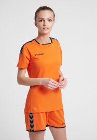 Orange atletisk trøje og shorts sæt med sorte accenter. Trøjen har en rund hals og logo. Shortsene har et chevron mønster på siderne.