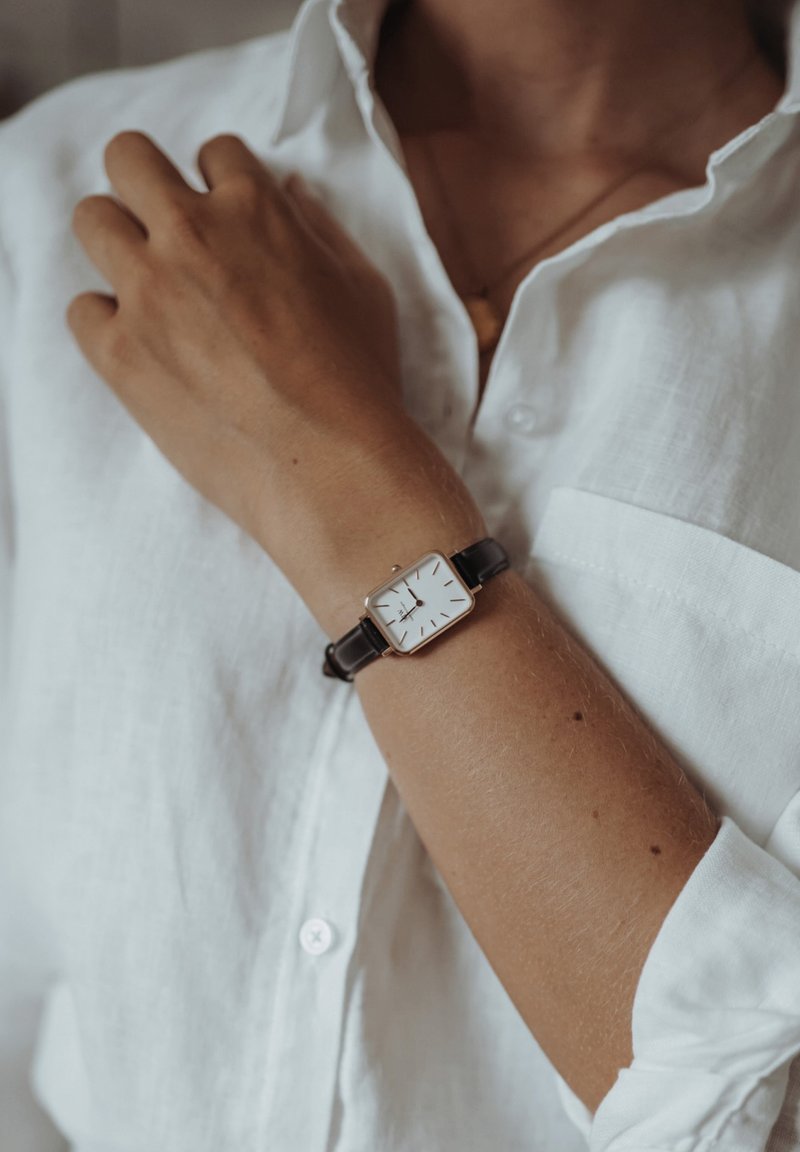 Square white watch with a gold frame and black leather band, worn on a wrist, accompanied by a white linen shirt.