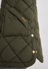 Olijfgroene gewatteerde vest met een grote zak aan de linkerkant, gouden hardware accenten en een subtiel logo nabij de zak.