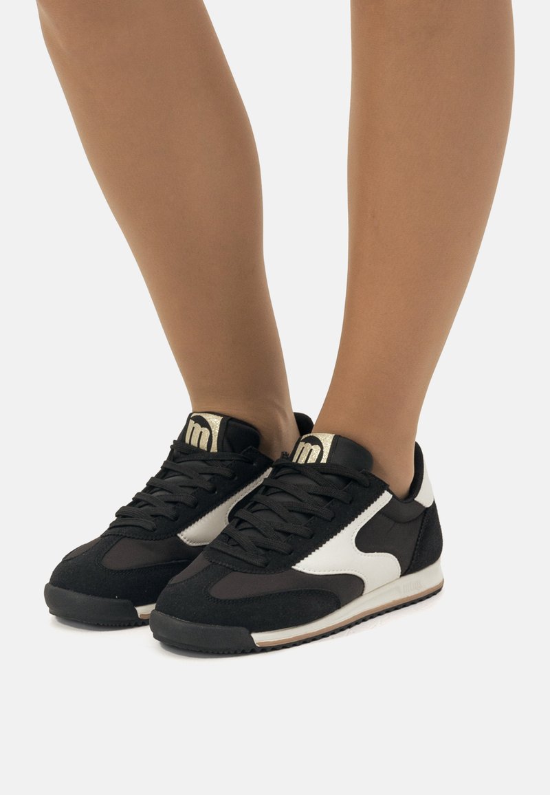 Scarpe sportive in bianco e nero con una tomaia in suede e sintetico, punta rotonda, spesse stringhe nere e dettagli dorati sulla linguetta.